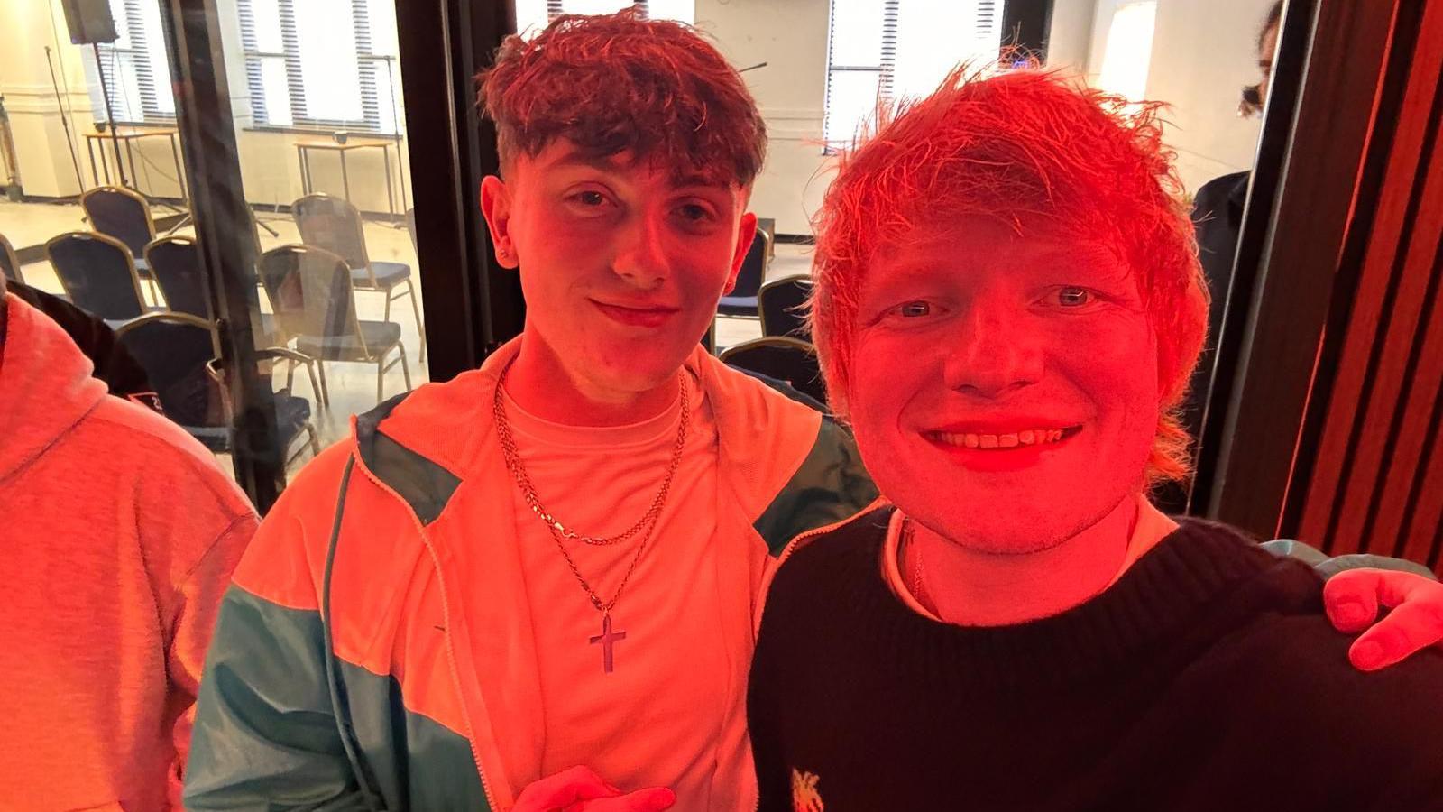 Ed Sheeran is posing for a selfie with a young man. They are in a studio with a red light shining. 