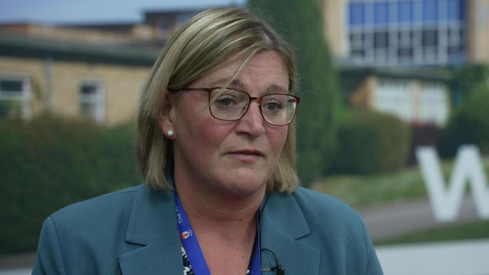 Kay Rimmer has shoulder length hair and is wearing glasses and a teal coloured suit jacket and black and white patterned blouse.