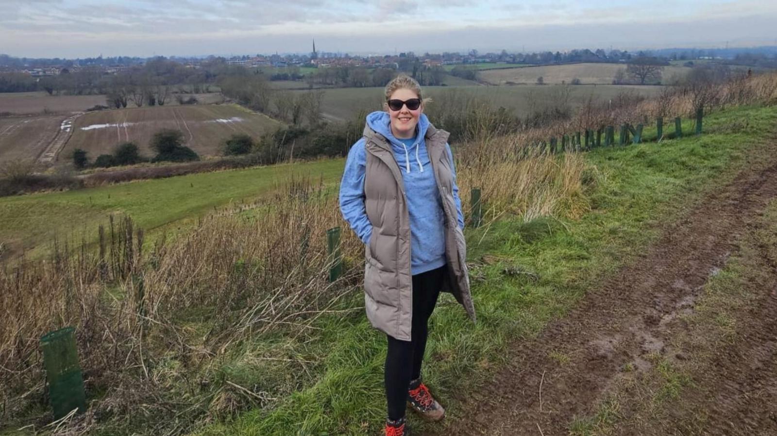 Tilly wears a light blue hoody and padded grey gilet. She is wearing black leggings and walking boots with red laces. She has black sunglasses on and her hair tied back, she is smiling at the camera and is on a muddy path with fields behind her.
