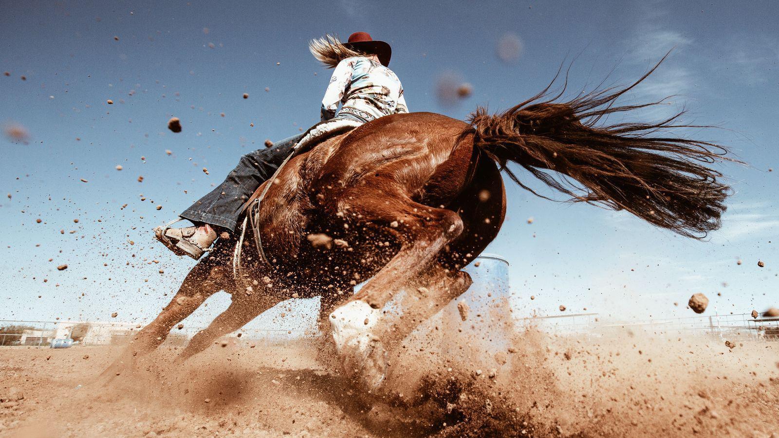 An action shot of a horse running through dirt and dust while a cowgirl rides it around a blue barrel.