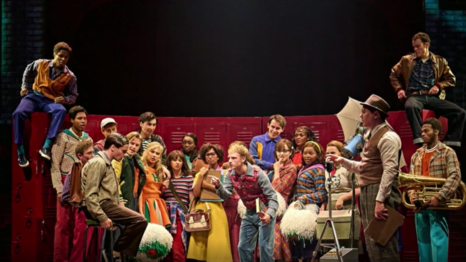 A full cast photo. 22 people are on stage stood next to a row of red lockers. All actors are staring to the left focusing in on one man. All of the people are wearing patterned clothes including striped tops, checked shirts and bright dresses.  