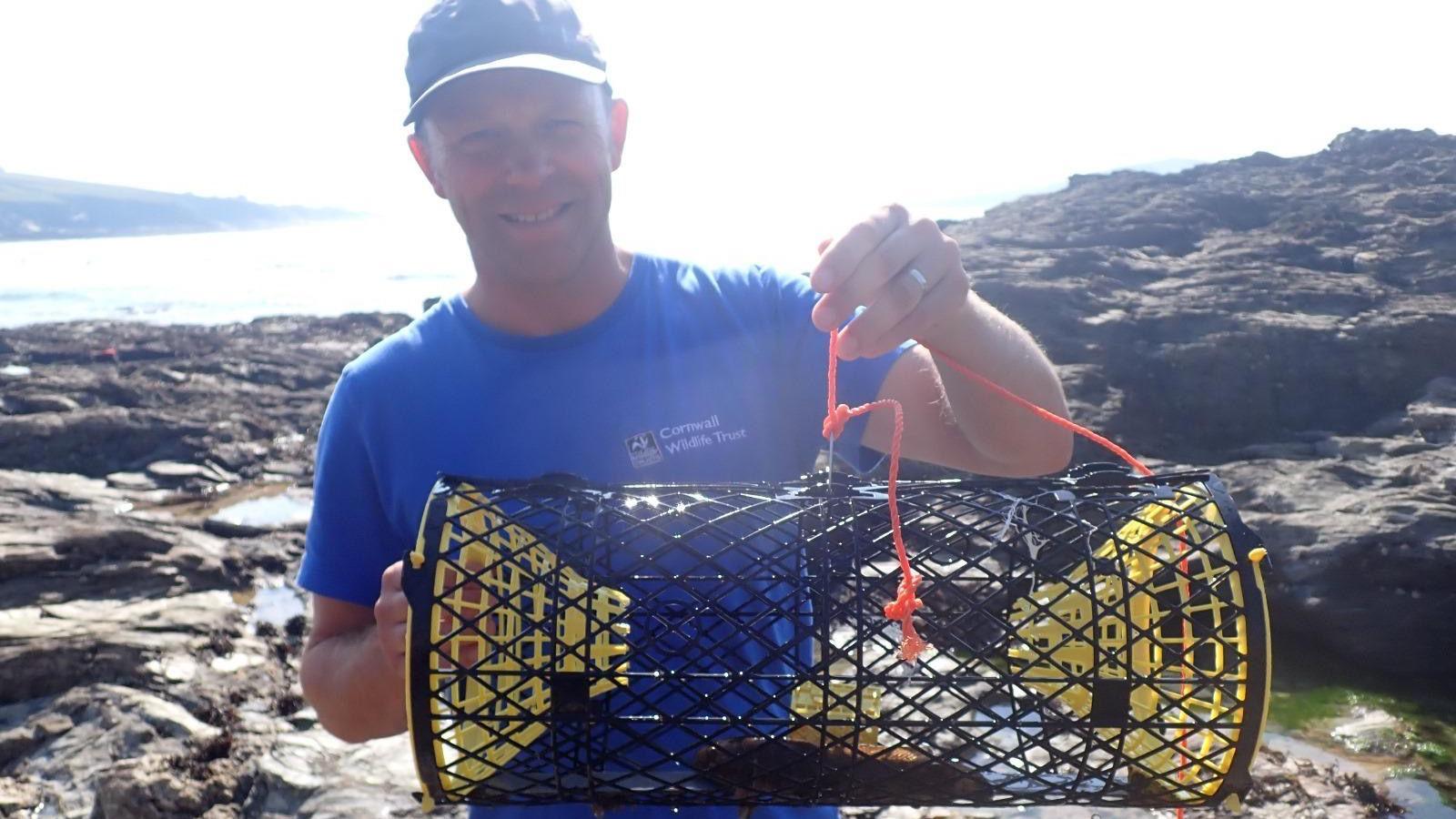 Matt Slater stands facing front wearing a blue hat and a blue T-shirt branded with Cornwall Wildlife Trust. He holds in front of him an oblong cage with what appears to be a lobster inside.