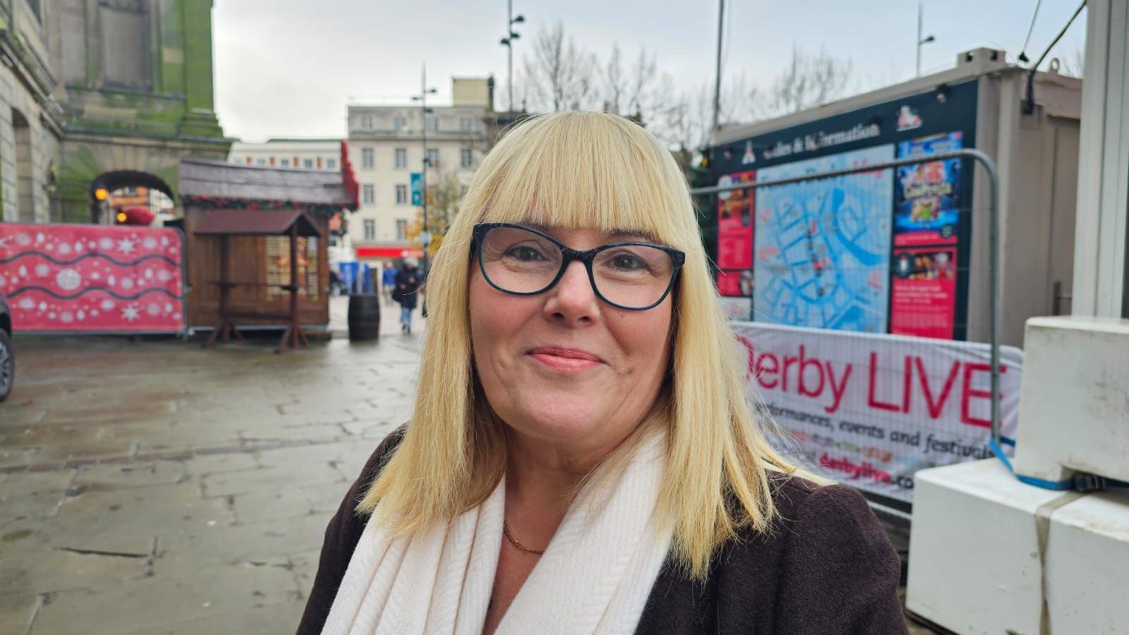 Carol smiles at the camera, she has blonde hair with a fringe, wears glasses and a white scarf. 