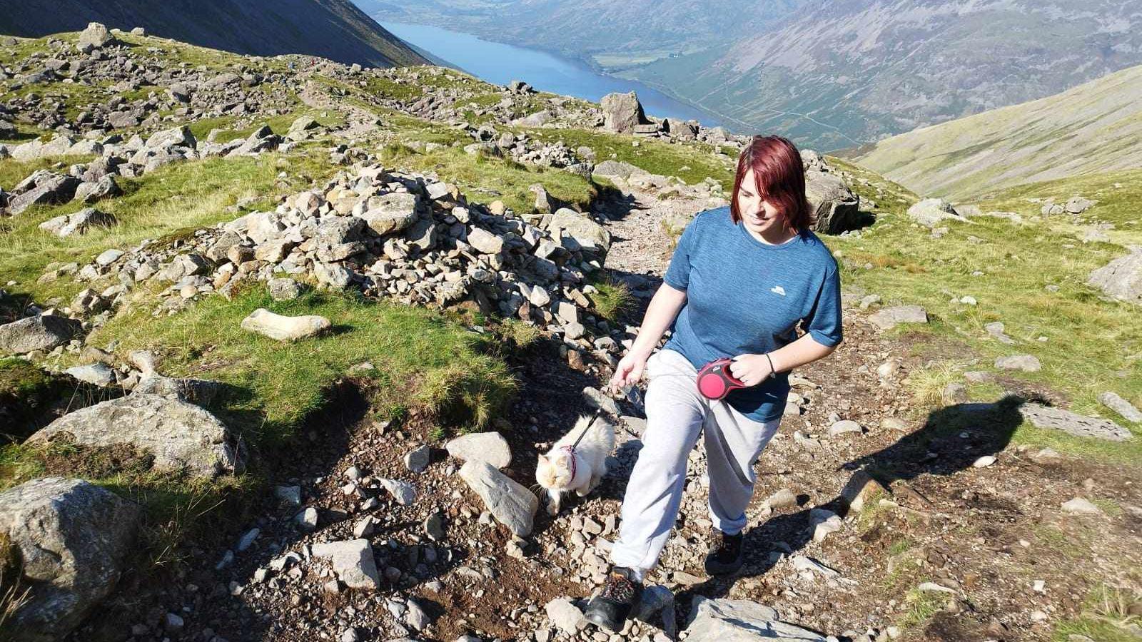 A woman in sports clothes and shoes walking with a cat on a lead on a mountain path. It's a sunny day. A lake could be seen behind them.