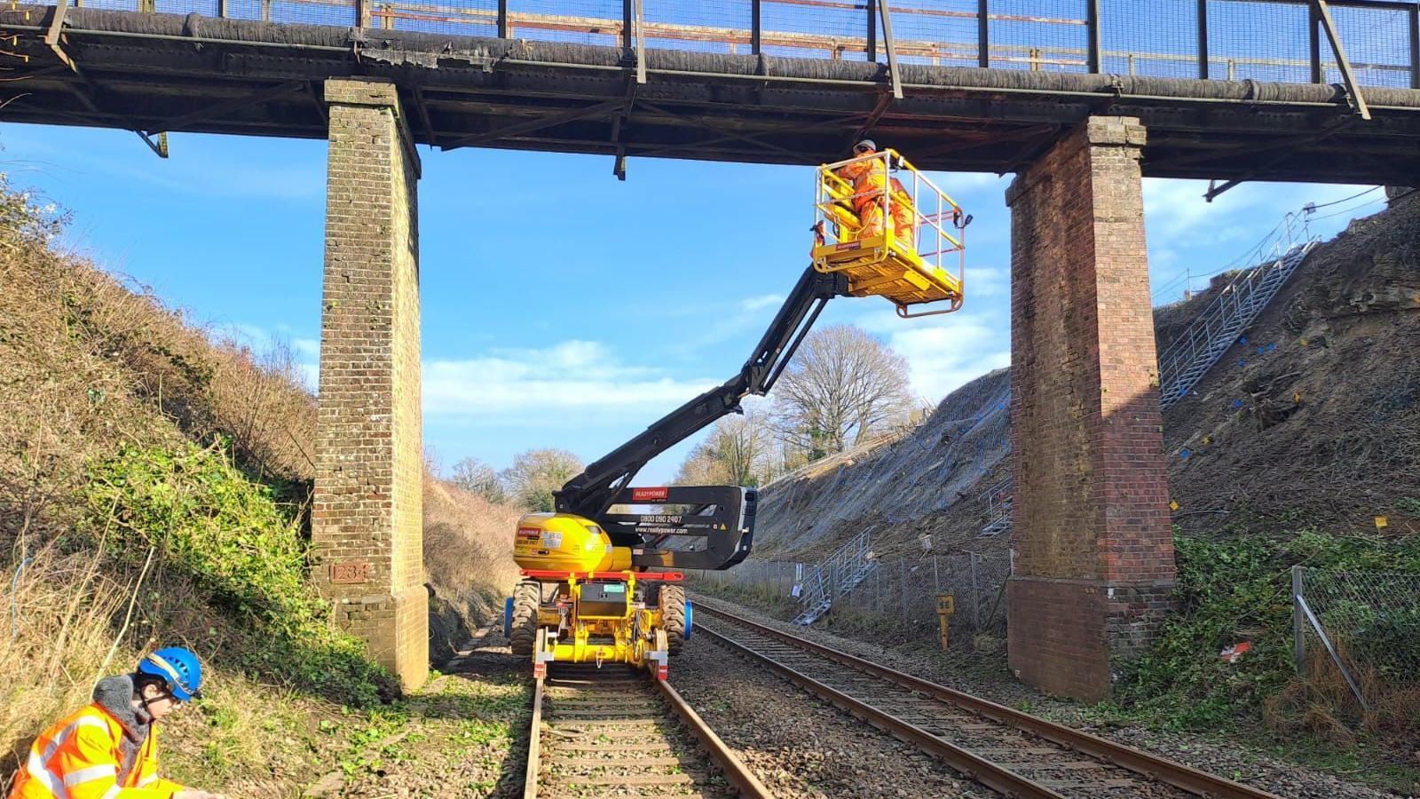 A yellow cherry picker is used to inspect a bridge over the rail line between Guildford and Gatwick Airport. A worker in orange hi-vis and a blue safety helmet sits at the side of the track.