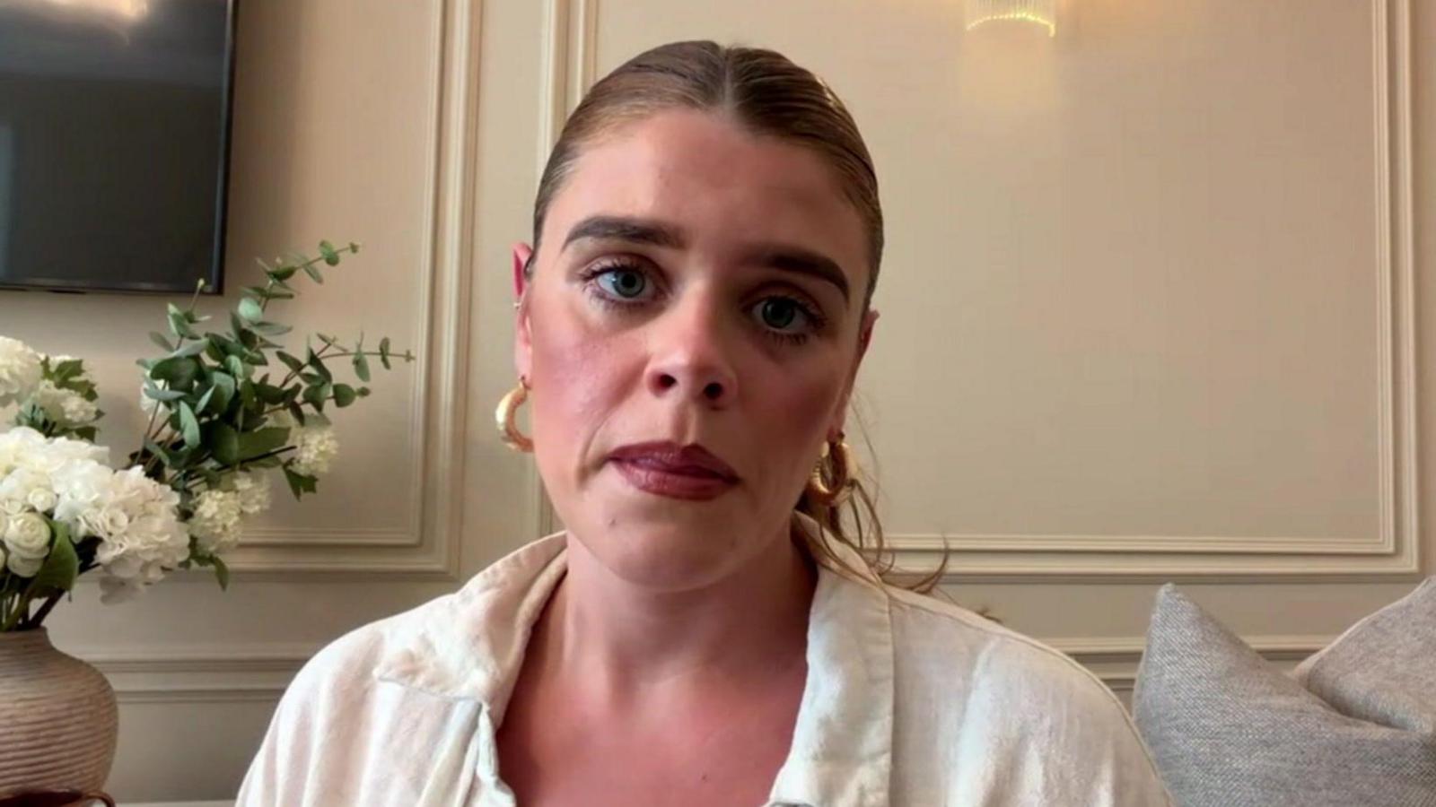 A woman with blonde hair scraped back into a pony-tail, red lipstick, gold earrings and a white linen shirt looks into the camera with a serious expression. 