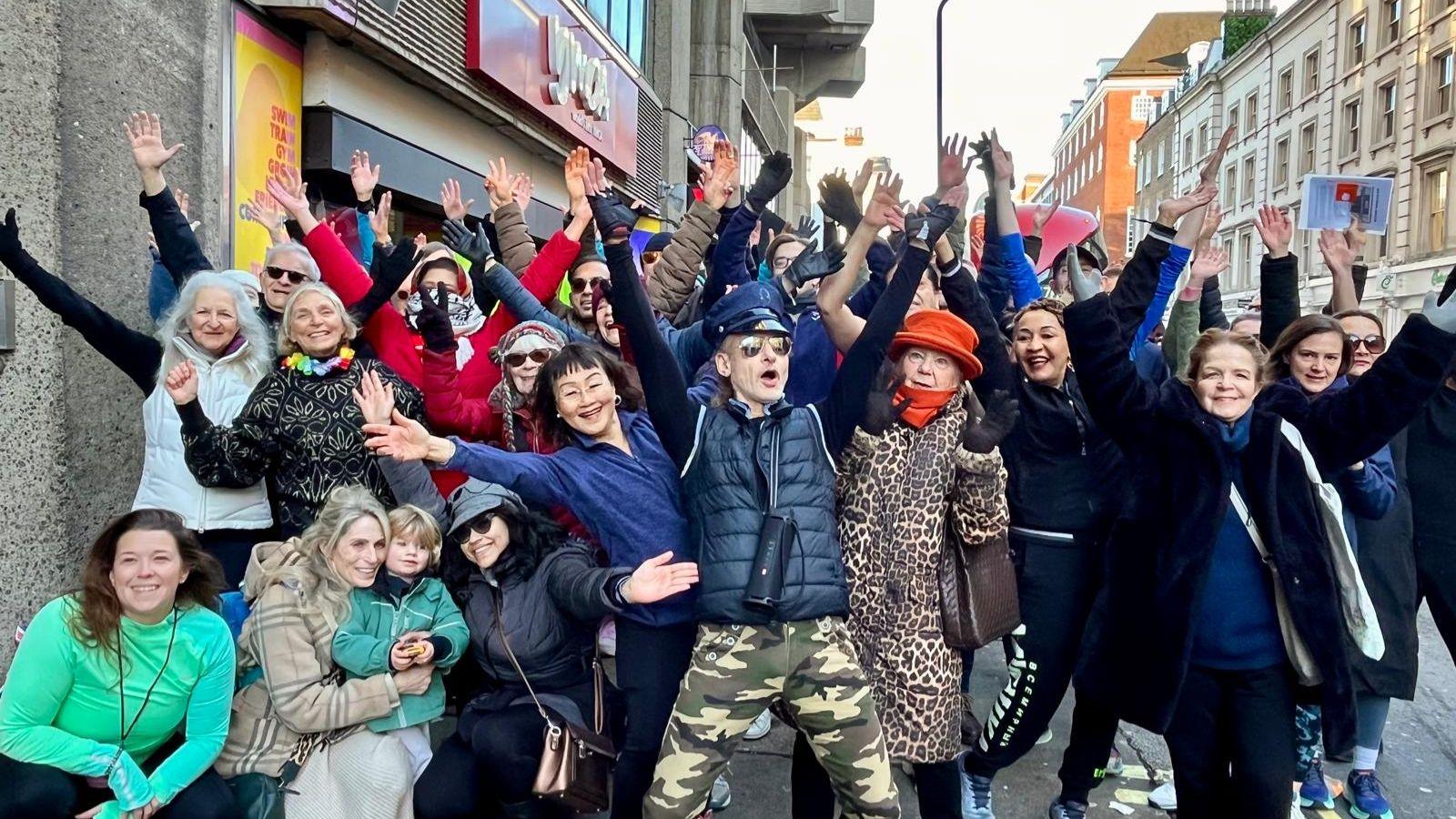 A group of 20 to 30 people standing with their arms up and smiling outside the YMCA. One man in the centre is wearing aviator sunglasses, camo trousers, a dark blue gilet and blue top, with a pilot's hat on.