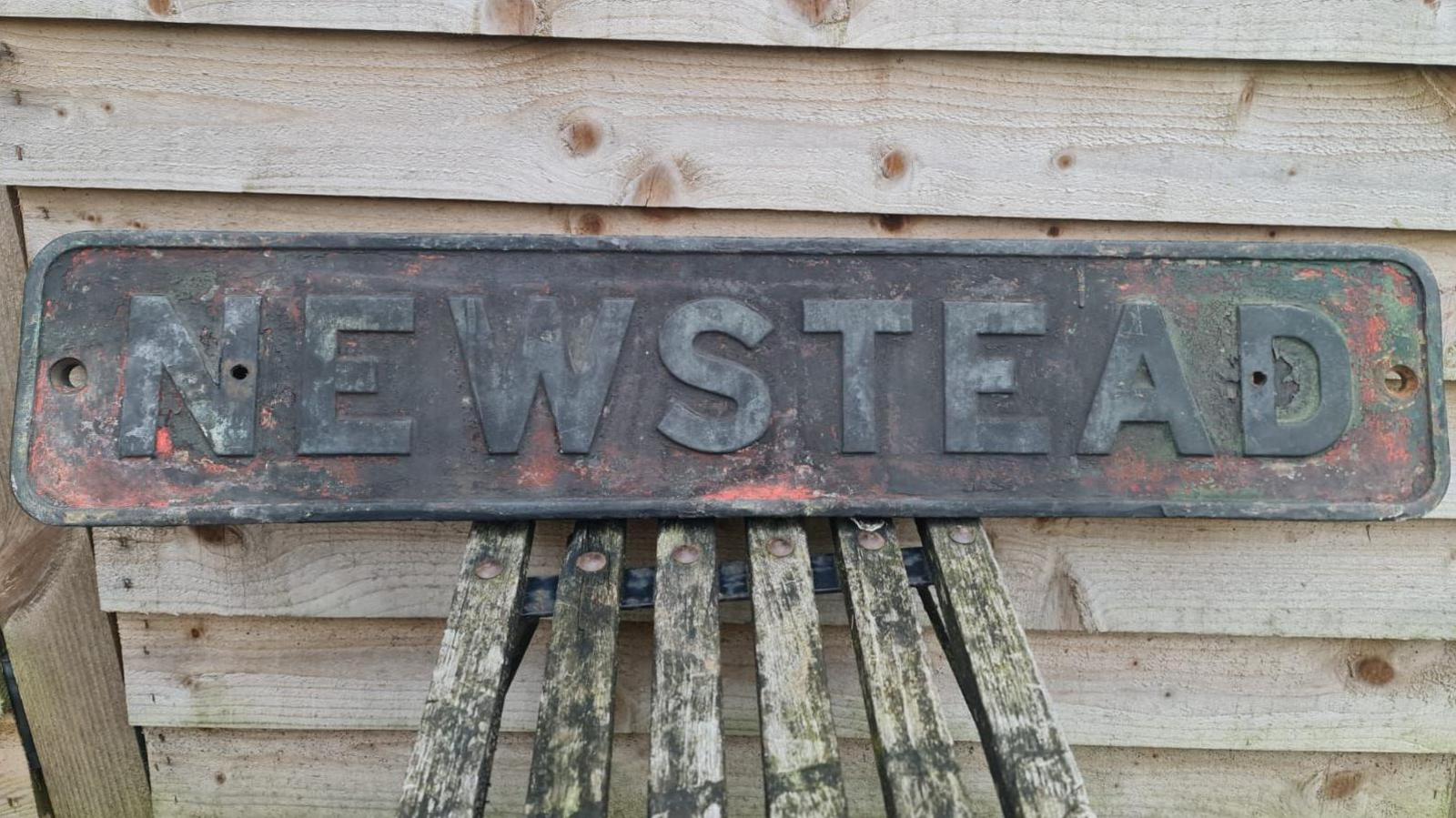 A brass name plate bearing the word NEWSTEAD in need of a lick of paint, leaned up against a wooden shed