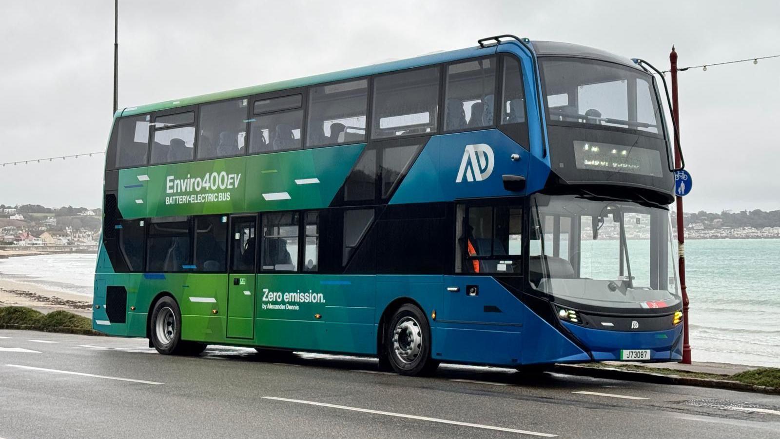 A modern, green and blue double-decker bus drives along a road next to a calm bay.