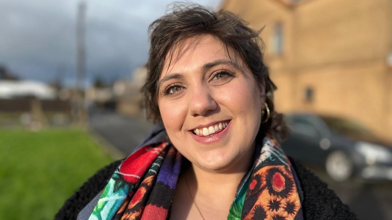 A woman smiles at the camera, she has short brown hair and is wearing a colourful scarf