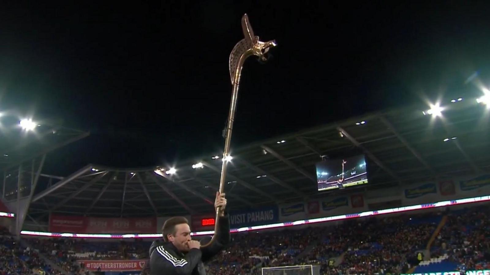 A man stands on a football pitch playing a long, bronze horn.