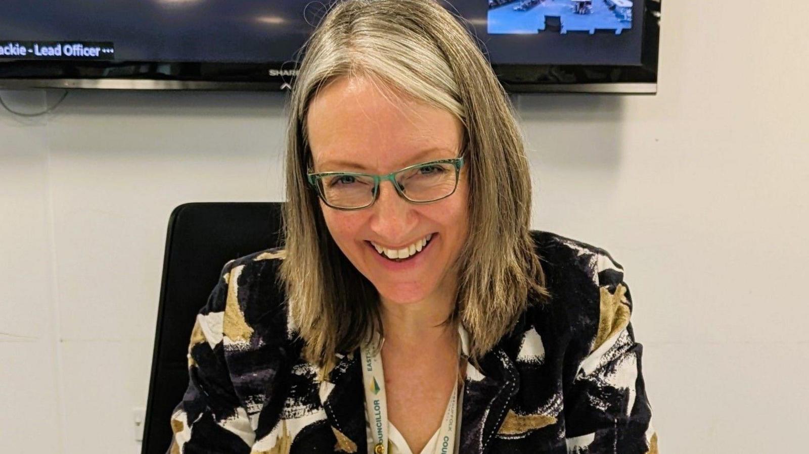 Caroline Topping sitting down and wearing a multi-coloured jacket and shirt. She is smiling and wearing glasses. 