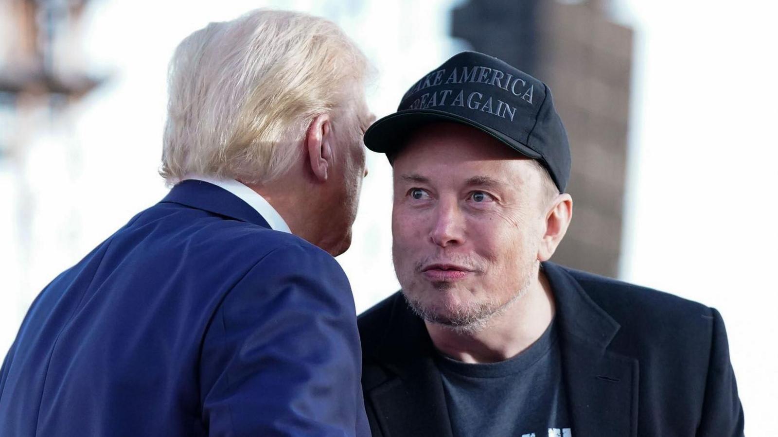 Tesla and SpaceX CEO Elon Musk speaks with former president Donald Trump during a campaign event in Pennsylvania. Musk wears a black 'Make America Great Again' hat and black blazer while Trump wears a blue suit.