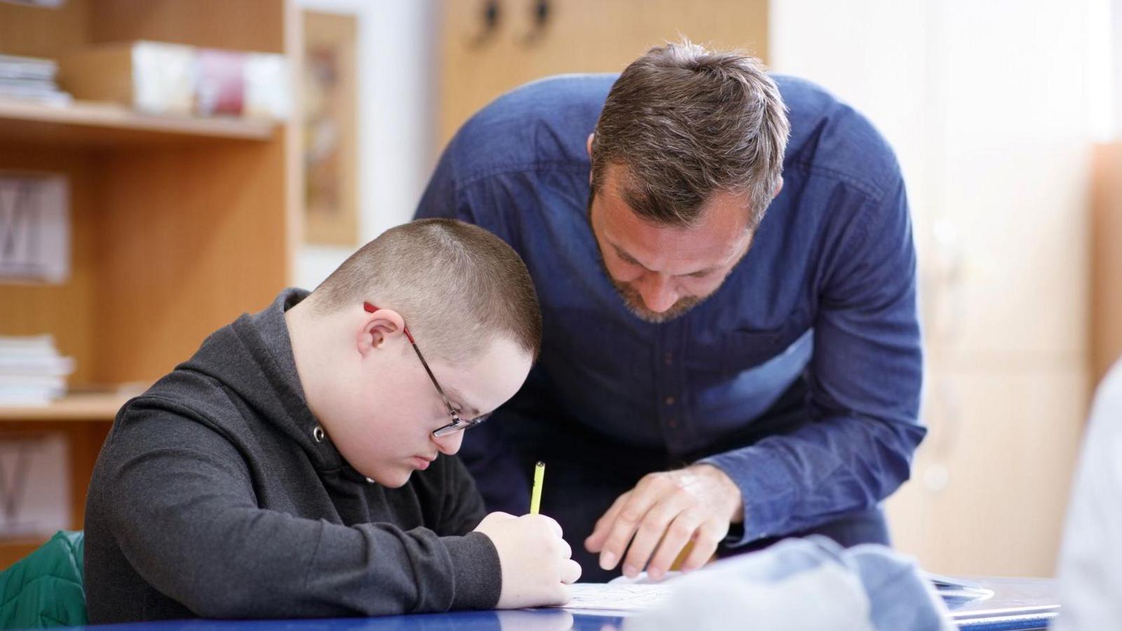 A male teacher wearing a dark denim blue shirt supports a student who is drawing on a page. The male student is wearing a dark grey hoodie.