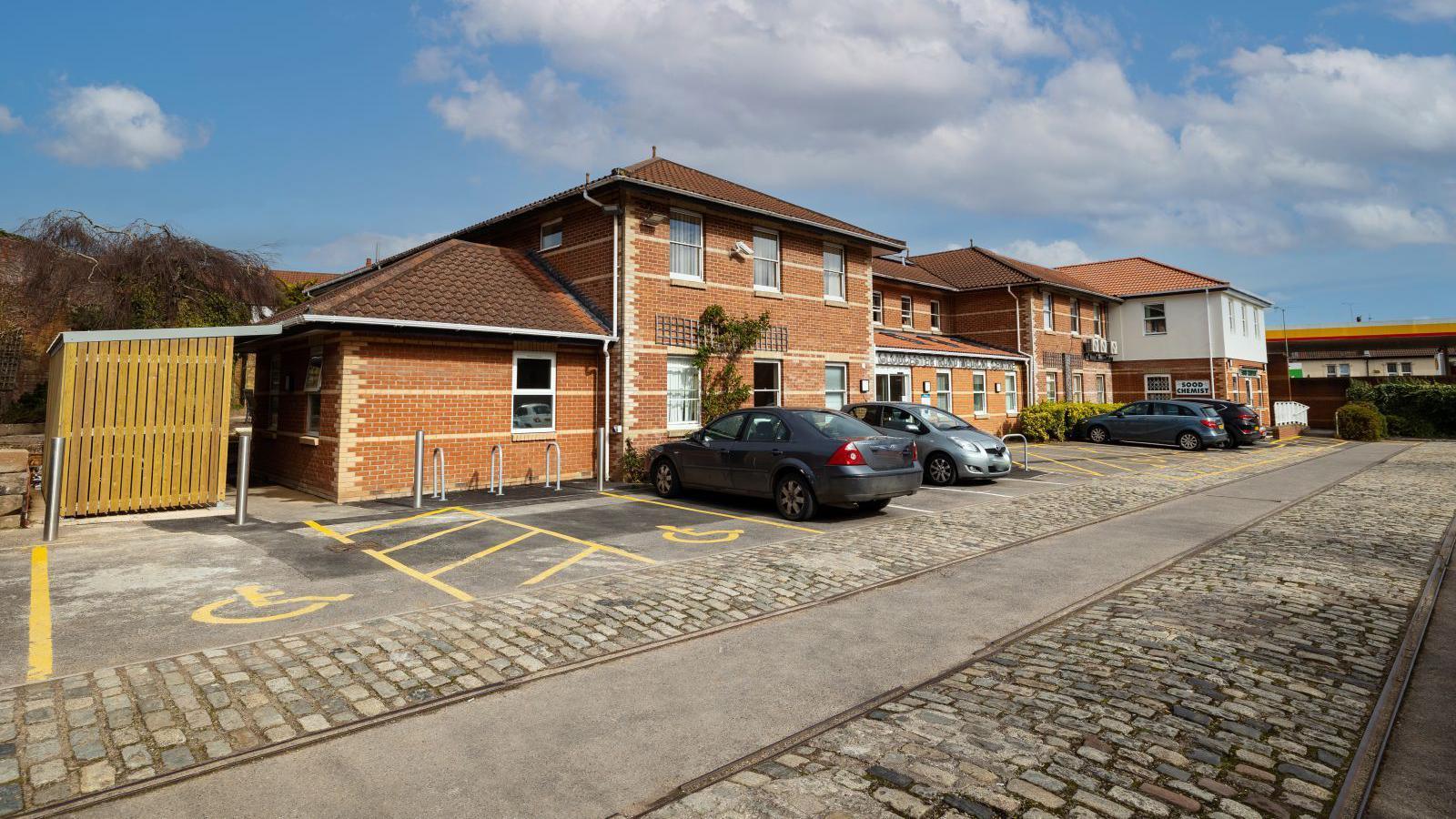 The Gloucester Road Medical Centre car park on a sunny day, facing towards the brick building and disabled car parking spaces. There are two tram lines running across the picture with concrete in between them and cobbled stones beside them. 