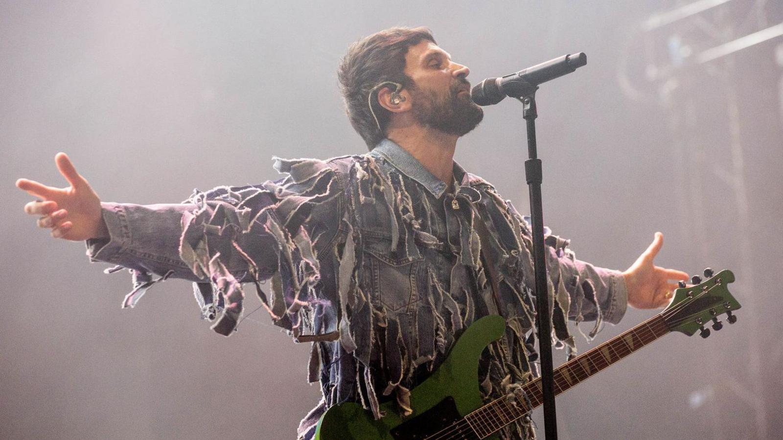 A man stands in front of a microphone with his arms spread apart as he looks up at the ceiling. He is wearing a shredded denim jacket and has a guitar slung over his shoulder.