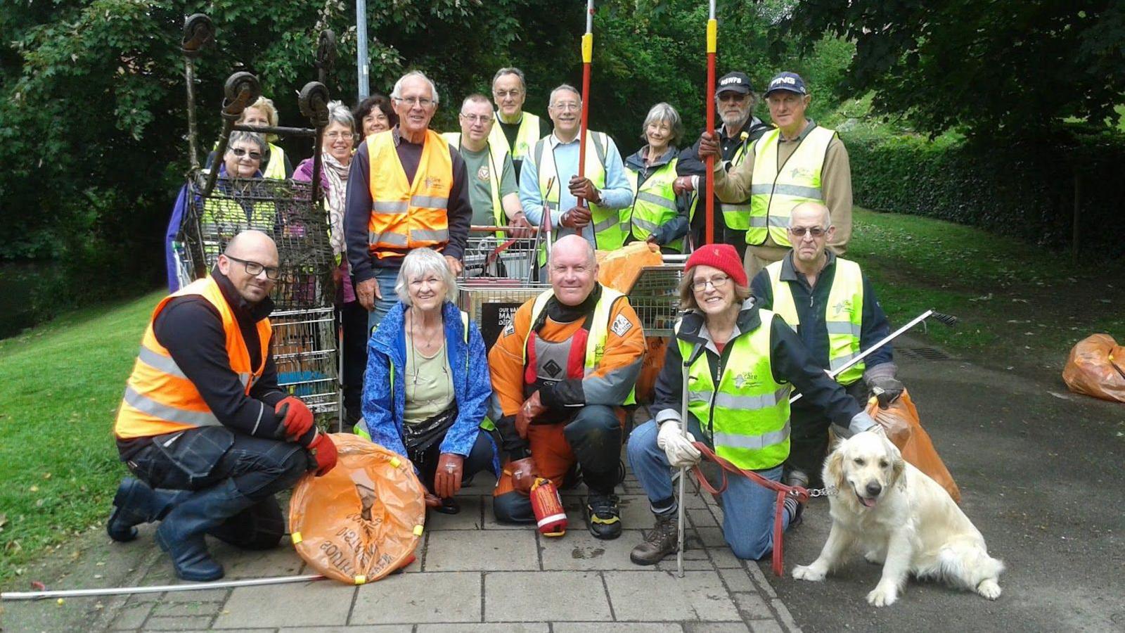 A group of litter pickers wearing blue, yellow and orange high vis vests, with some standing on top of a wall and others sitting below it. They are holding litter picks and bags and there is also a golden retriever.