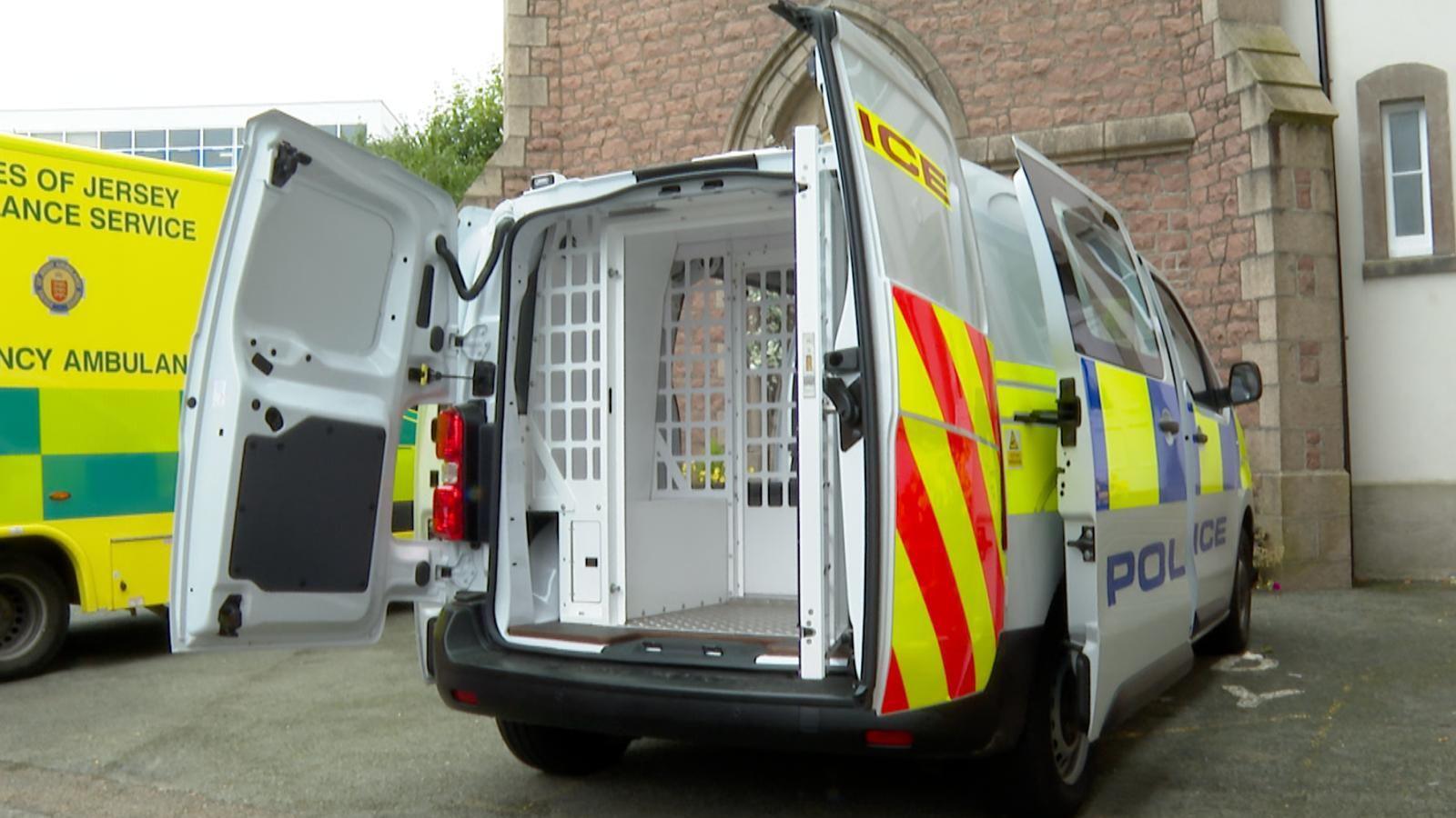 A white police van parked with its backdoors opened to show a small prison within the back, parked in front of a granite next to a yellow ambulance on the left