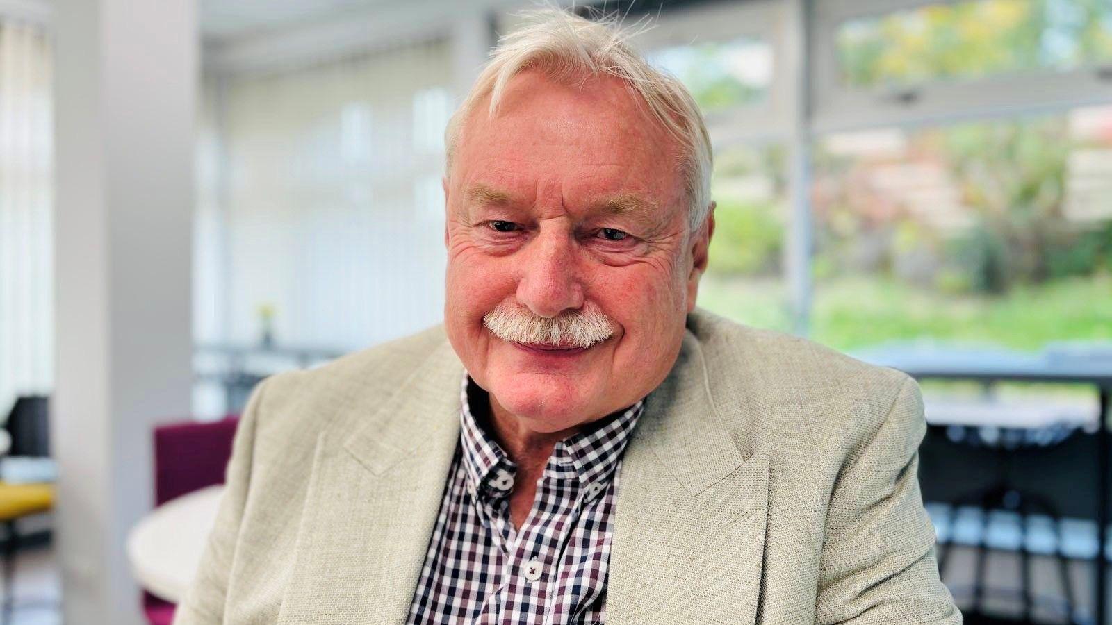 Prof Wayne Powell, man smiling, with grey moustache, wearing checked shirt and beige jacket, with window in background