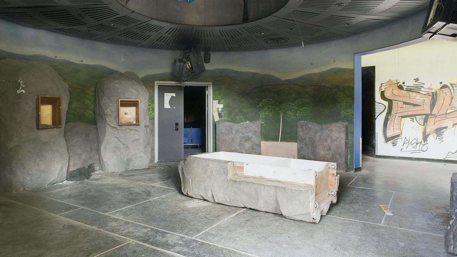 The inside of the park. The room has large stones around the circumference which look run down and damaged. There is a broken door and graffiti on the walls.