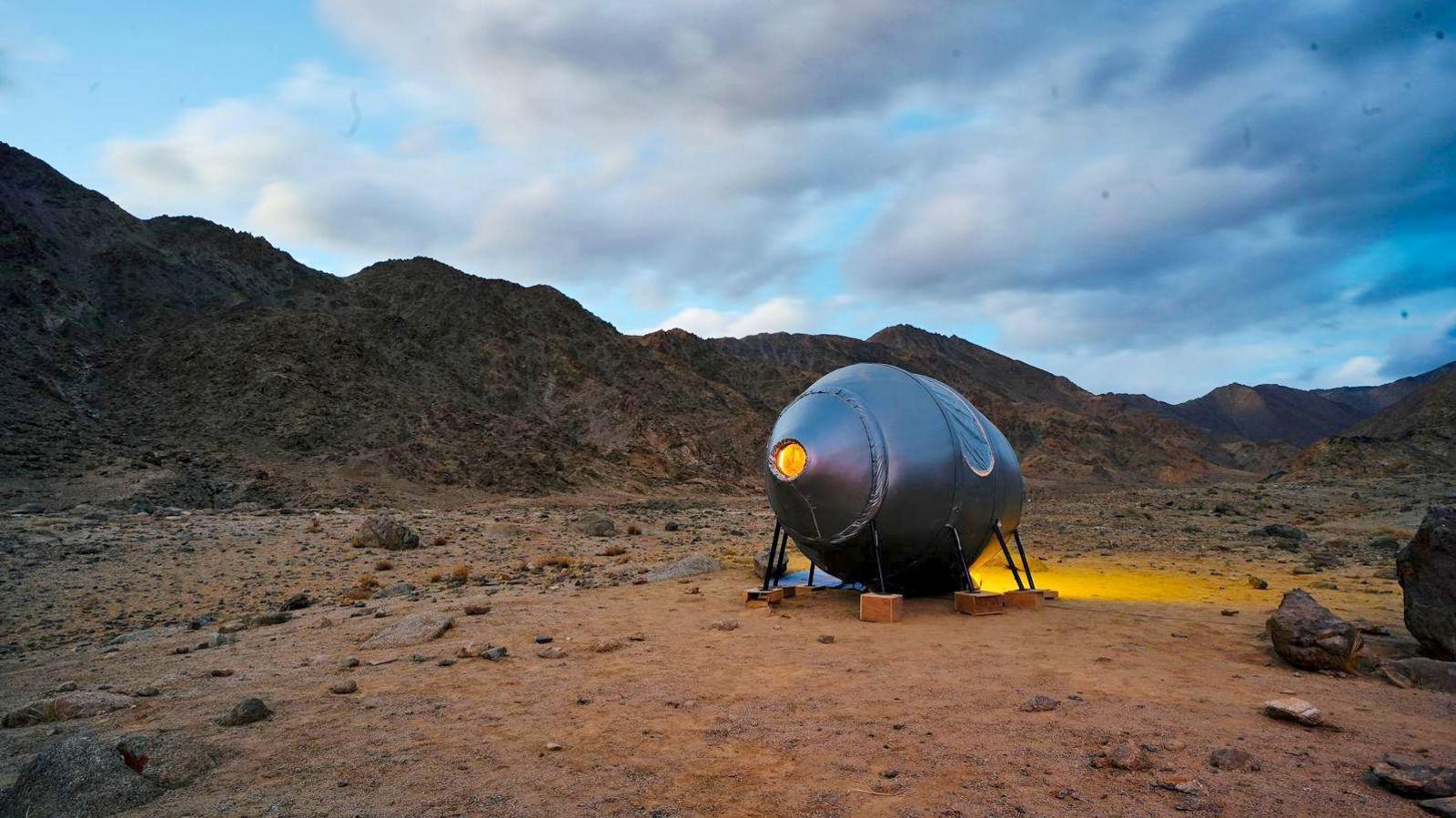 A circular pod is seen parked in the mountains of Ladakh, it is sliver in colour and held up with little pole structures held in place with plywood boxes, with the browny-red backdrop of the Ladakh mountains in the background and some clouds in a dark sky.