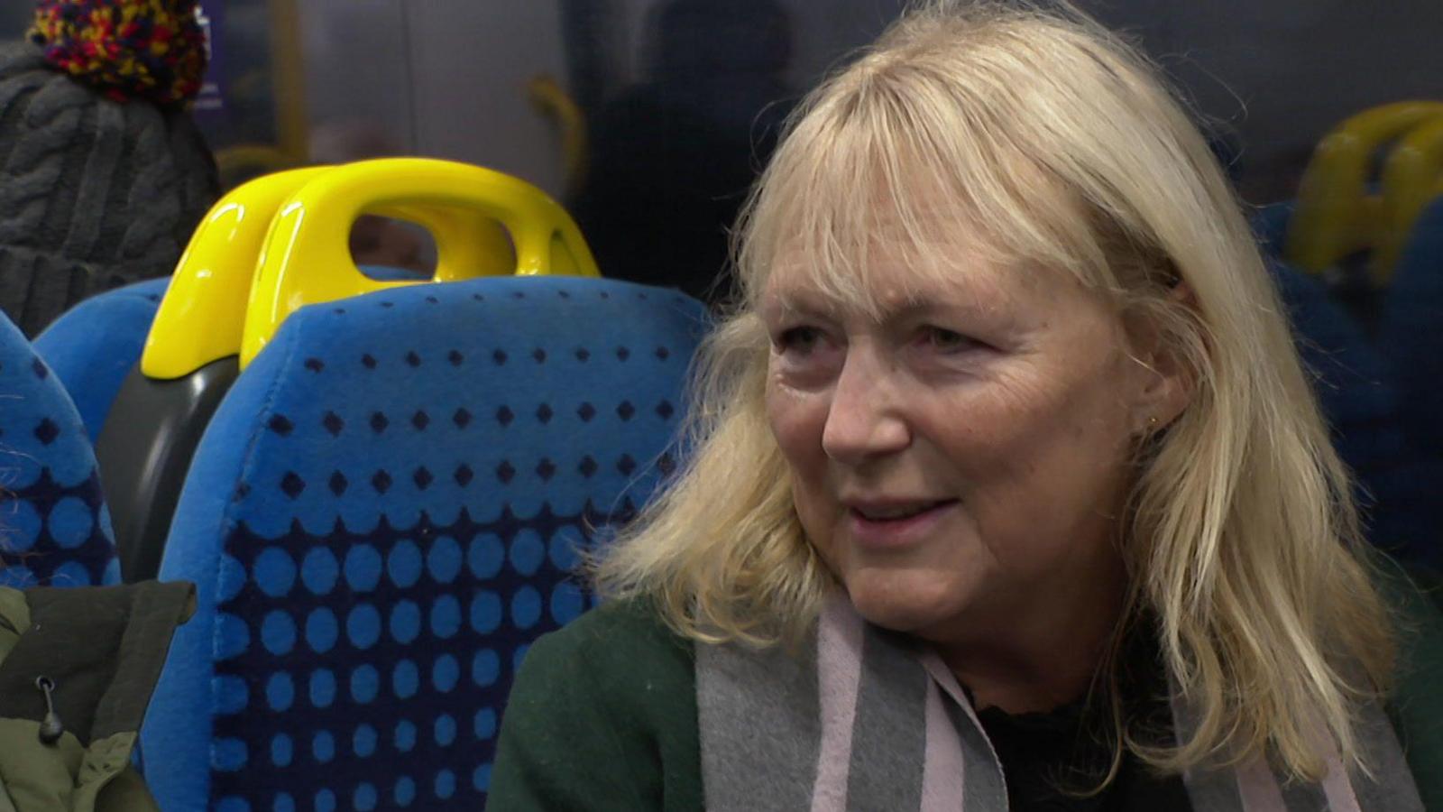 Clare Harland, a woman with blonde hair wearing a green coat, sits on a train