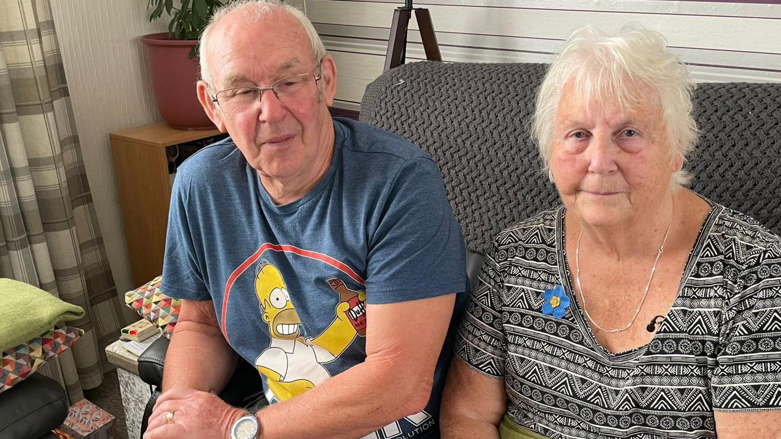 A man and a woman sitting on a brown-coloured sofa. The man has short grey hair and glasses and is holding his right arm with his left hand. The woman has light-coloured hair and is wearing a grey and white patterned dress and is looking at the camera.
