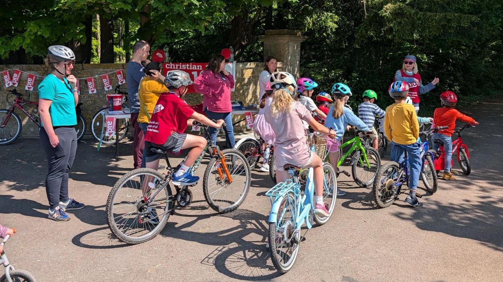A number of children riding colourful bicycles in dappled sunlight with Christian Aid bunting on a wall in the background.
