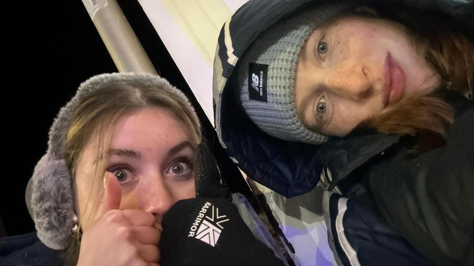 A selfie of two women, one wearing ear muffs, gloves and giving a thumbs up, and the other wearing a hat and a coat with her hood up. The photo is taken at night and there's a lamppost behind them.