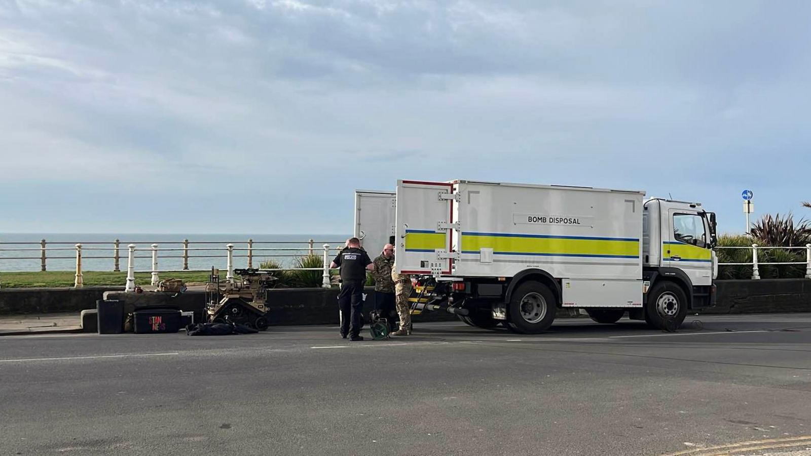 A bomb disposal van parked at the side of the road. The back doors are open and people are retrieving equipment from it.