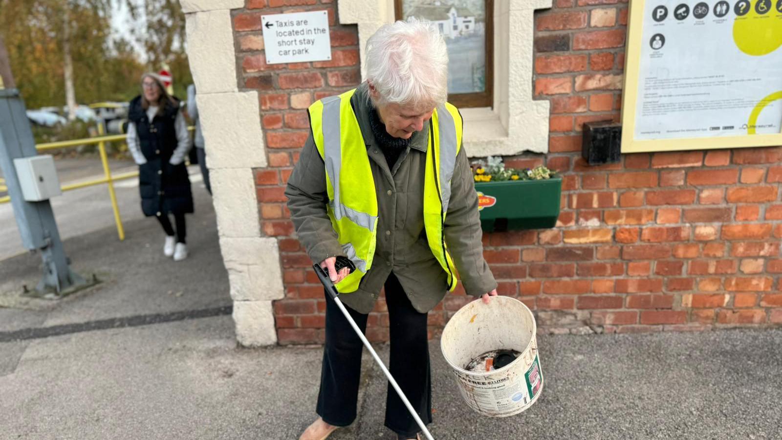 Margaret Walton, who has short white hair and wears a dark green coat with a high vis jacket, holds a litter picker and a plastic bucket