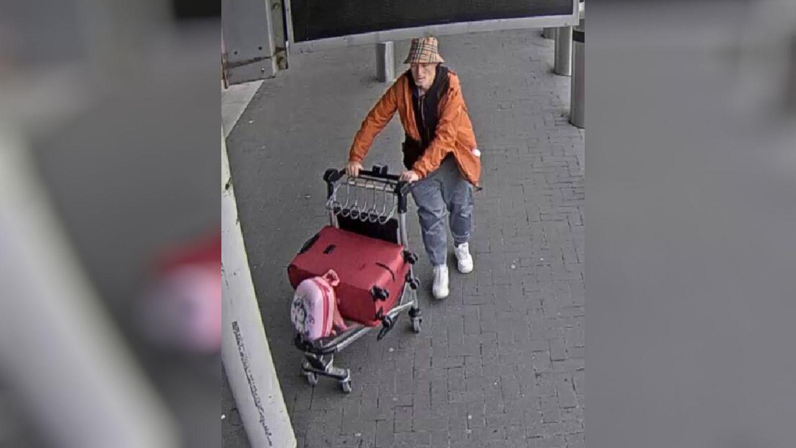 A man with a bucket hat and orange jacket leaves an airport wheeling a trolley with luggage on it. 