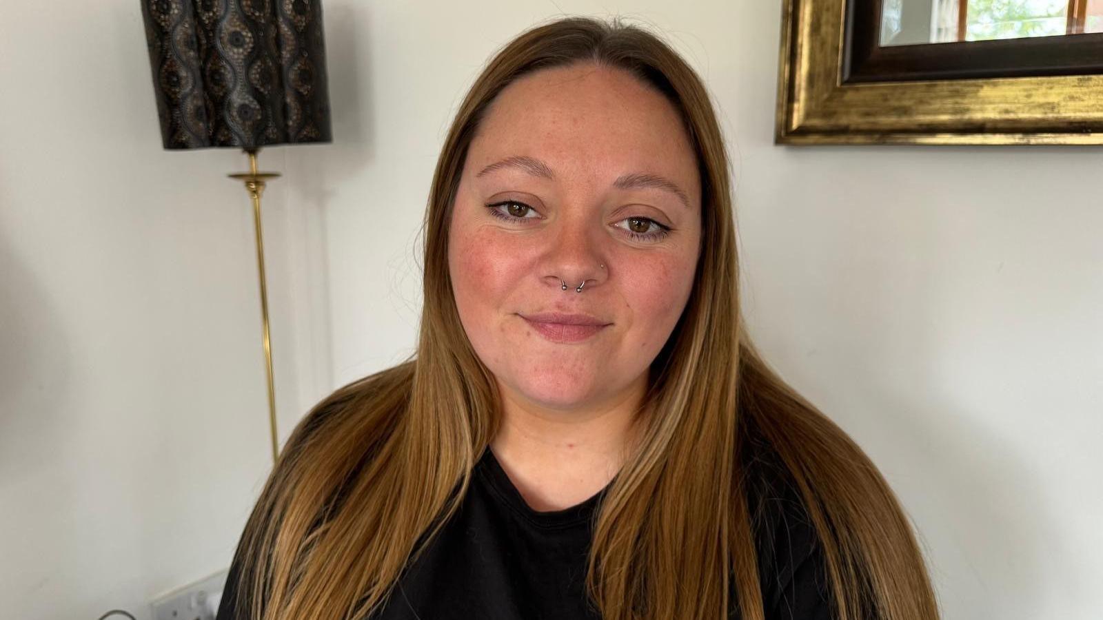 A woman with brown hair is looking into the camera. She has a septum piercing 