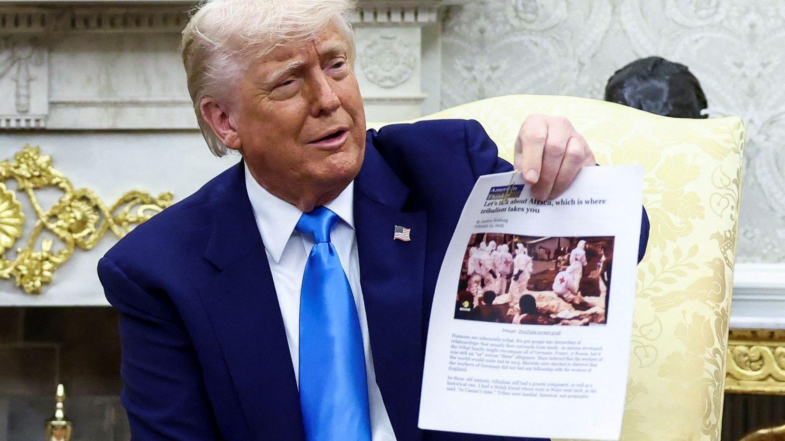 Donald Trump holding up a piece of paper with an article on it. An image of people in white medical suits can be seen. Trump is wearing a dark suit with a light tie and an American flag pin.