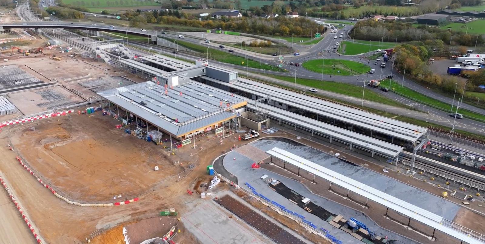 An ariel view of the construction site for Beaulieu Park station. A rail line runs through the middle of the site and to its left are silver roofs over the platforms and station building. Brown/orange earth is exposed on the left where excavations have taken place. In the distances are roads and a roundabout, and some landscaped trees.