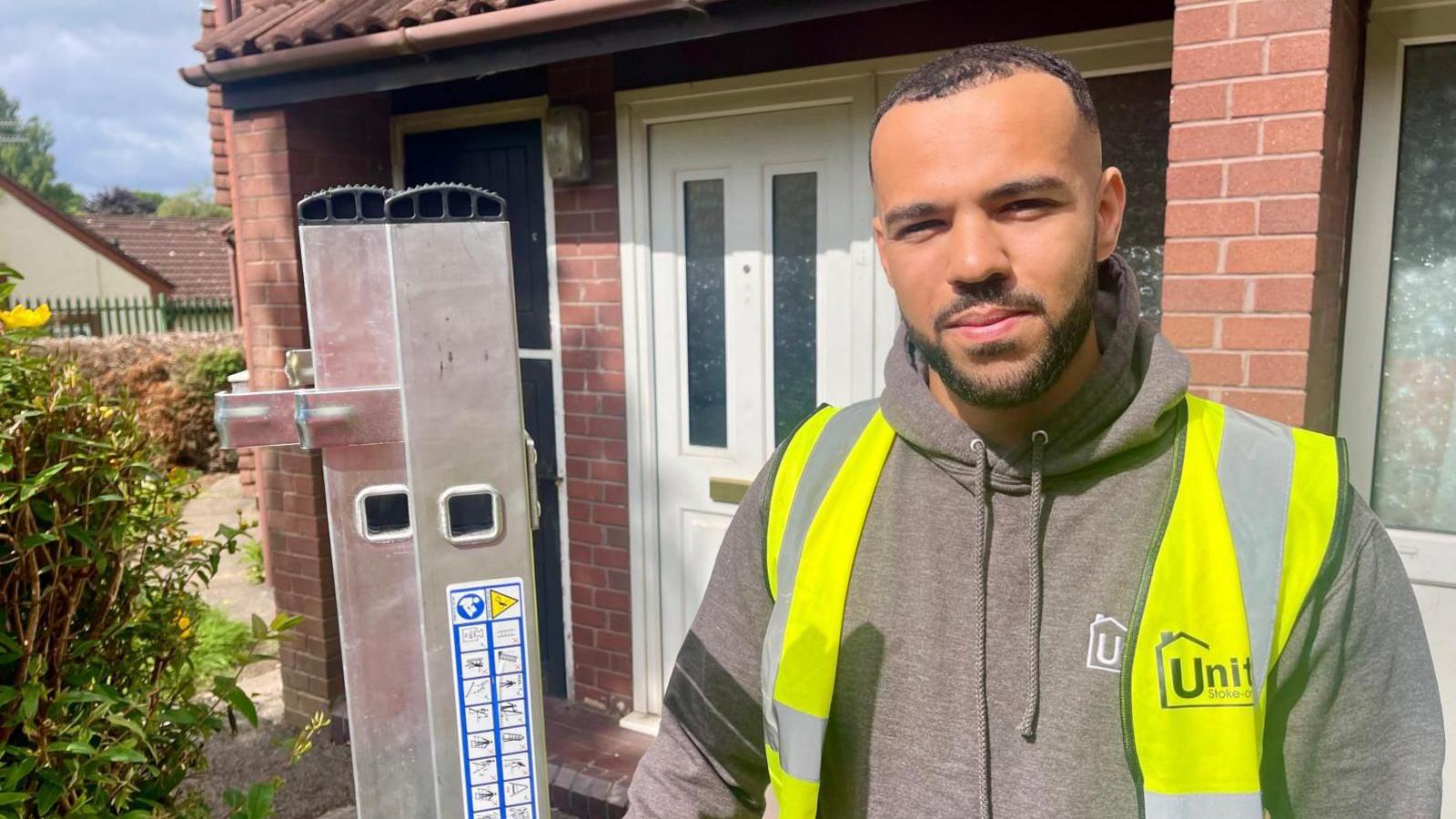 A man in a hi-vis vest and light brown hoodie holds a ladder outside a semi-detached house. It is a sunny day with bushes to the left of the man.