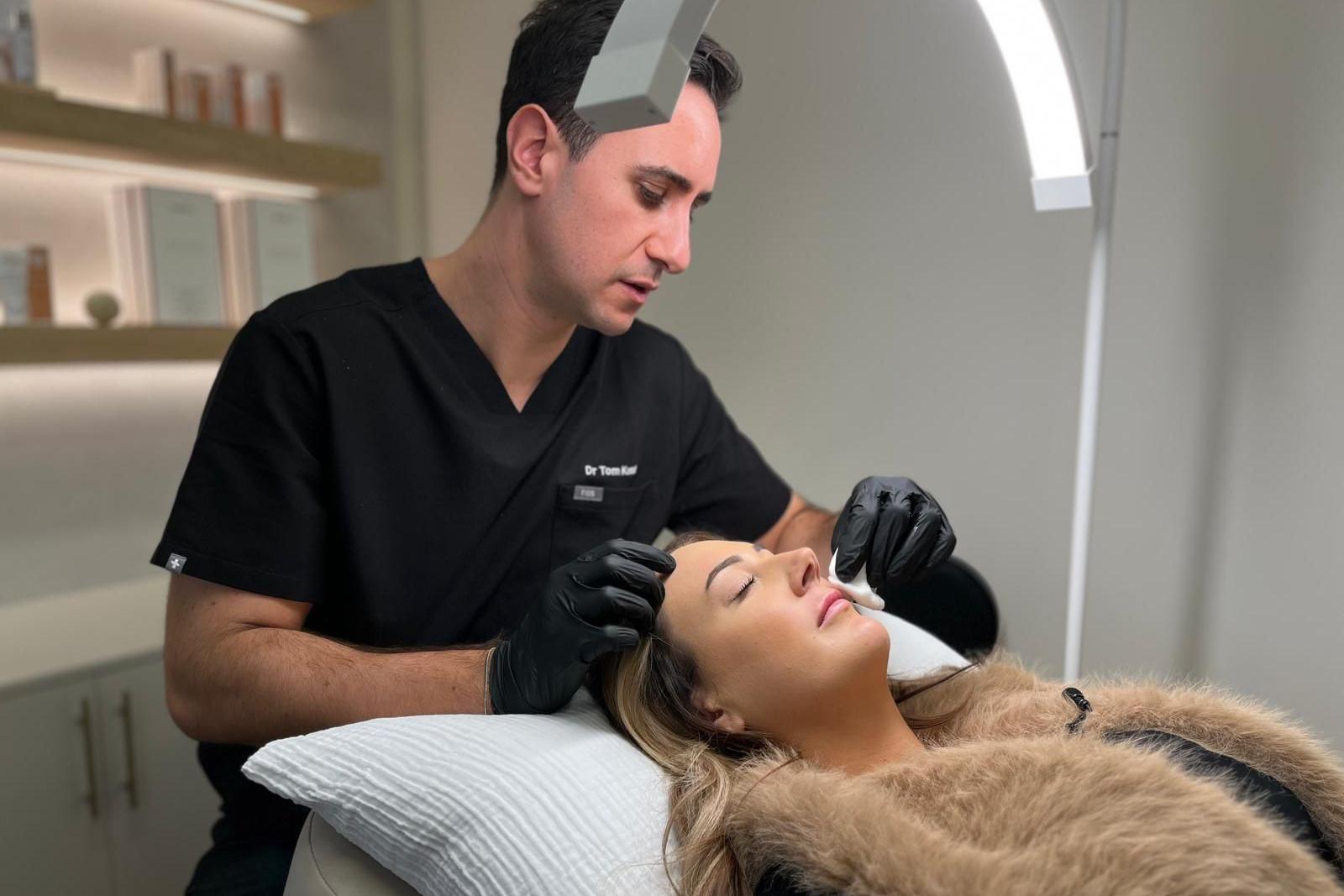 A woman is lying on a treatment bed with her eyes closed. A light is shining on her face and Dr Tom Kamal is wiping her lips after injecting her upper lip with botox. He is sat to the left of the frame slightly leaning over her. He is wearing black disposable gloves and a black scrub-type top.