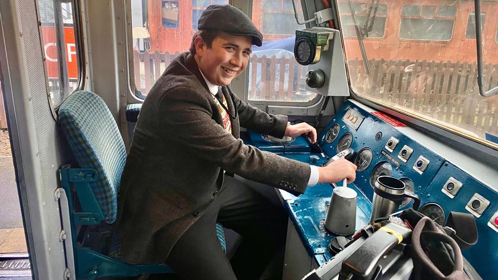 Logan is dressed in a flat cap and blazer - dressed like he's in the 1950s - sits in the front of a train.