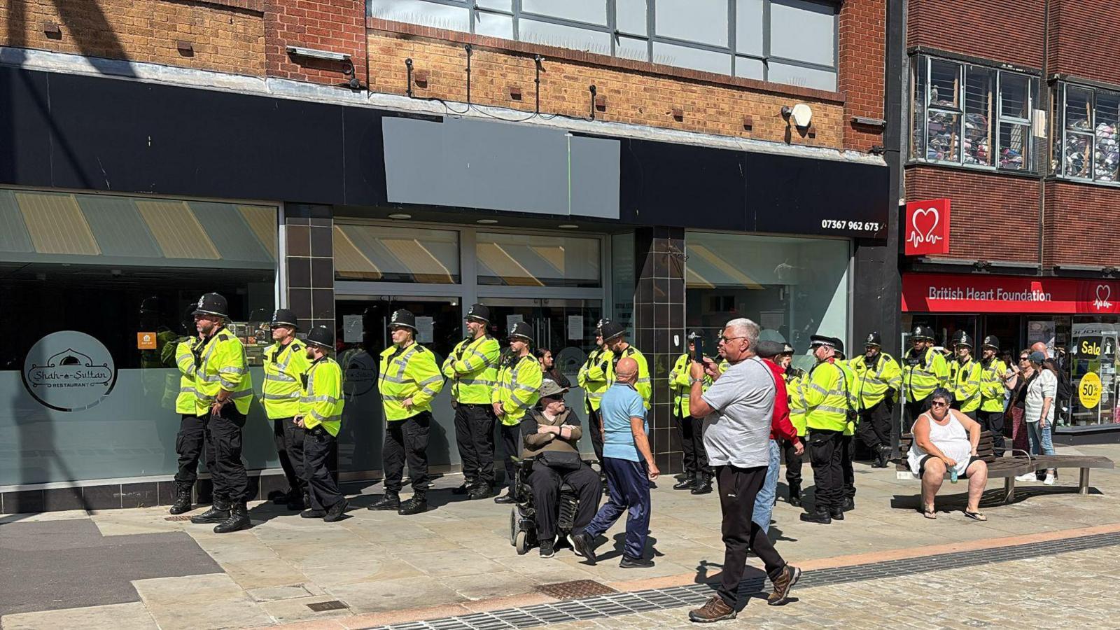 Police lined up before the protest took place. The officers are standing outside a restaurant and charity shop. They are wearing helmets and hi-vis jackets. Some members of the public are sitting and standing nearby.