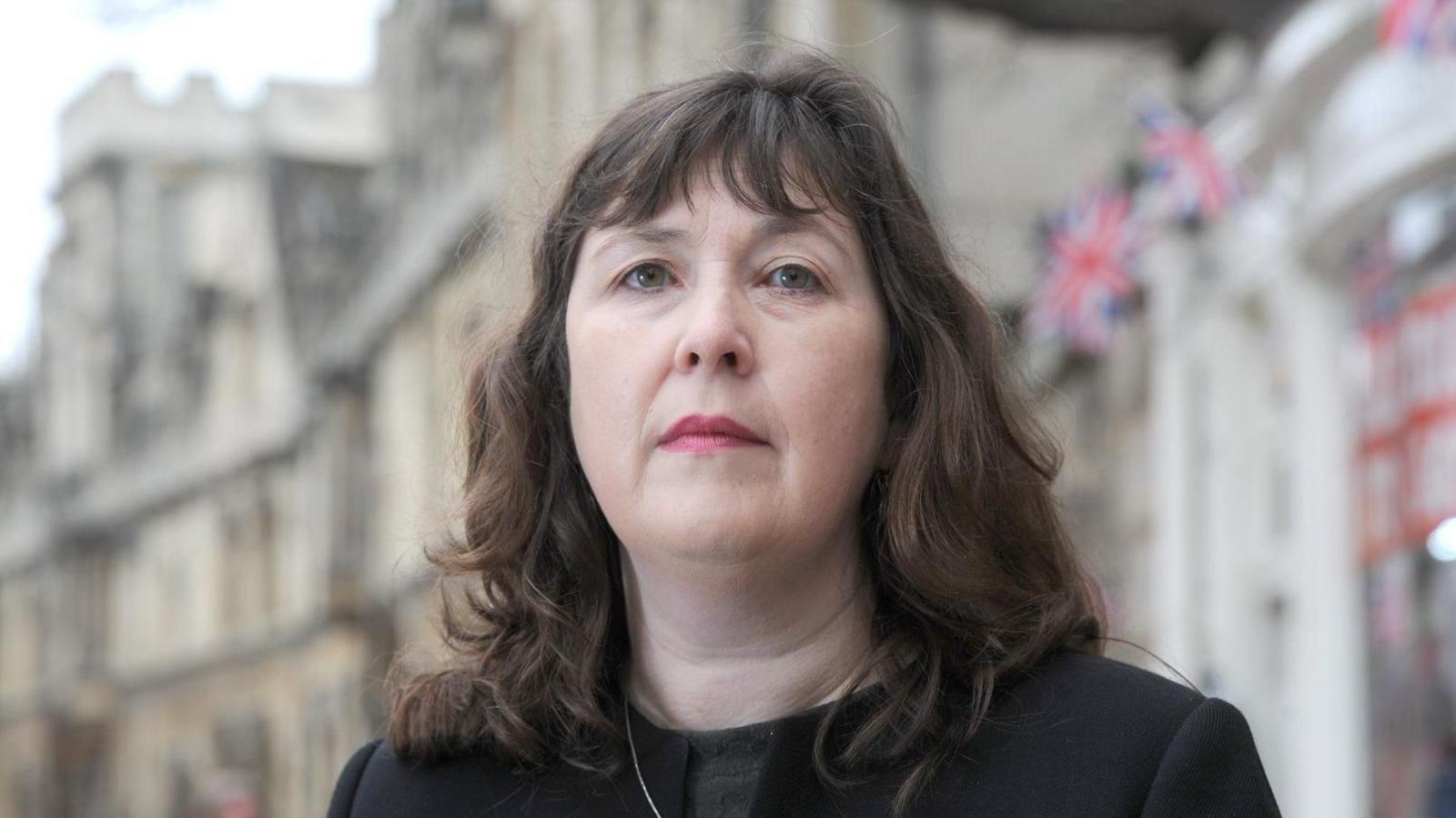 Susan Brown, who has shoulder length brown hair, wearing a black suit. She is standing in a street. Out of focus Union Jack flags are in the background.