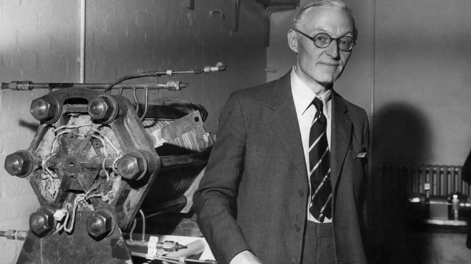 Tom Bacon is seen in an old black and white photograph standing next to the fuel cell he developed. He has receding fair hair, dark-rimmed round spectacles and is wearing a a dark suit, a white shirt and a striped tie