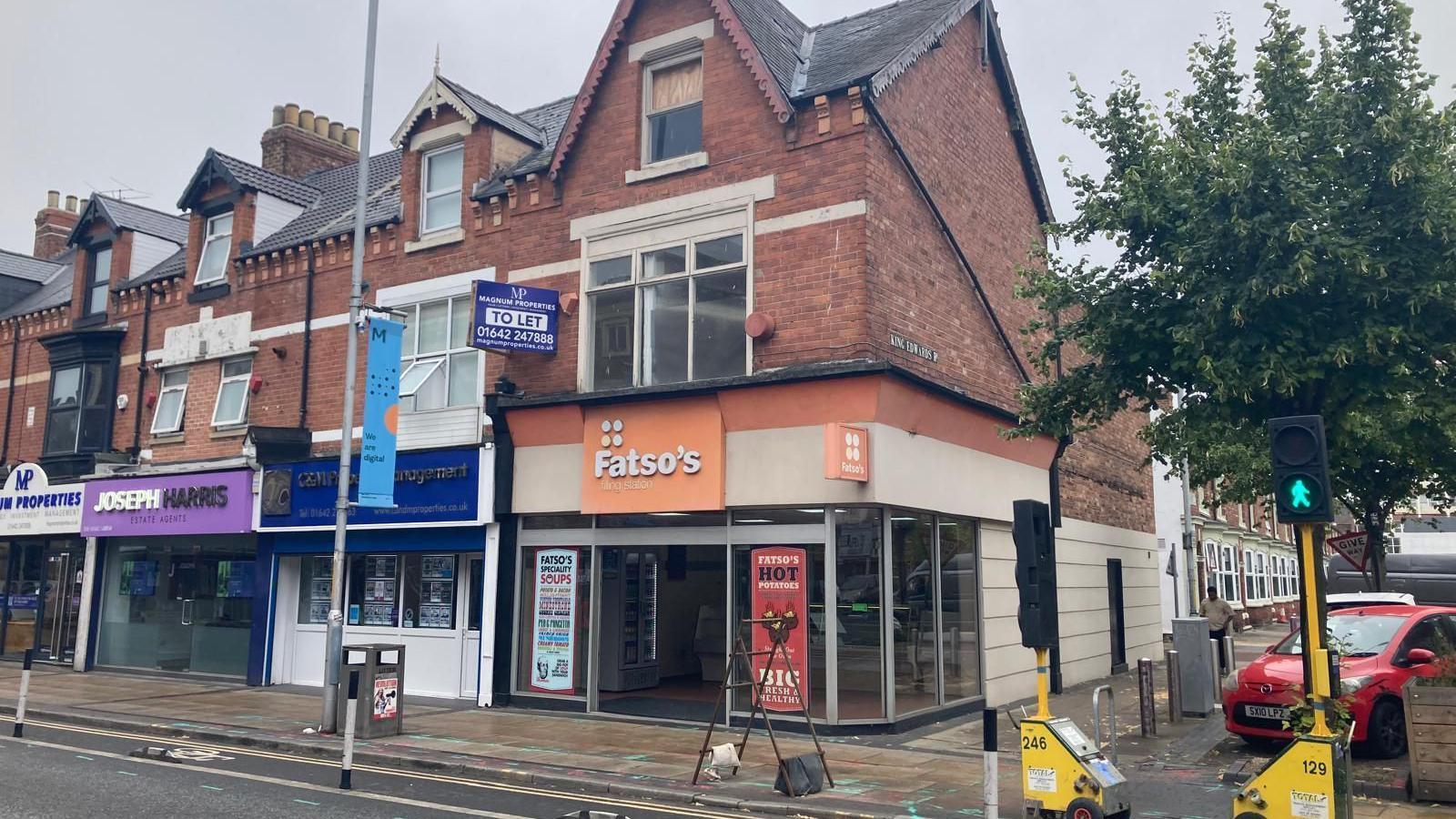 Fatsos located on Linthorpe Road. A building sits on the corner of a road, with large glass frontage and an orange sign over the entrance which reads ' Fatso's' in white lettering. It sits on a row of shops and a road is in front of it with temporary traffic lights. 