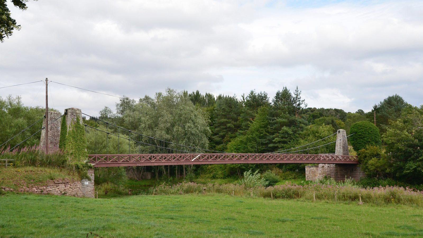 A side view of a suspension bridge - the river is not visible but it is surrounded by trees and green fields
