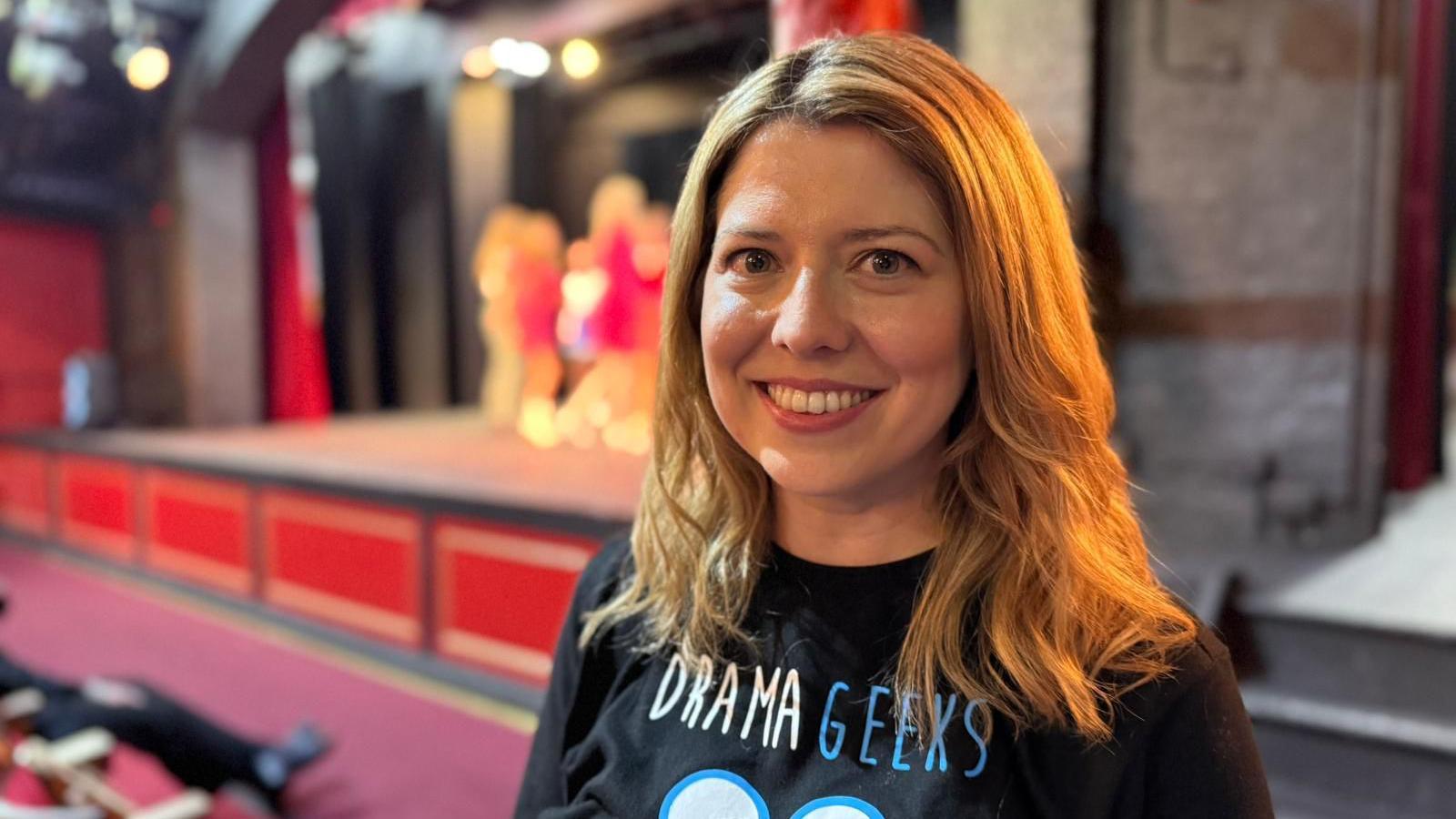 Charlotte Reid smiles at the camera. She has shoulder length blond hair and is wearing a black t-shirt with "Drama Geeks" written on it in white and blue letters. Behind her is a stage with several young people on it.