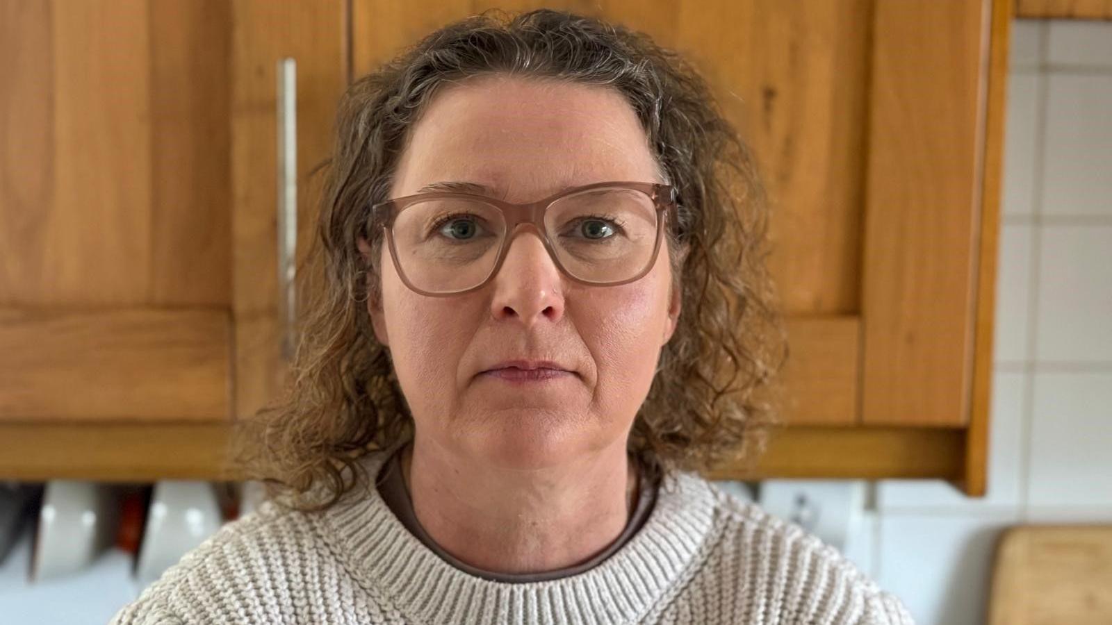 Victoria Fear qëndron në një kuzhinë me një dollap dhe tasa pas saj.