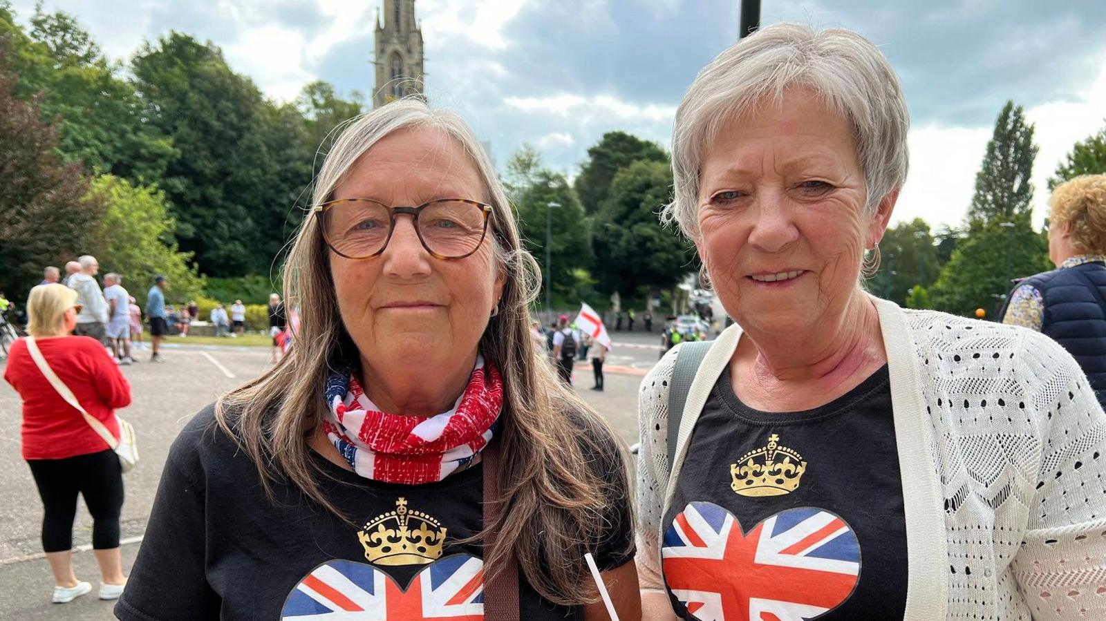 Two women, one with long greying hair and glasses and one with short grey hair, wearing t-shirts with heart-shaped union jacks on. They are smiling at the camera. Behind them is a car park with people milling around on it, some holding England flags.