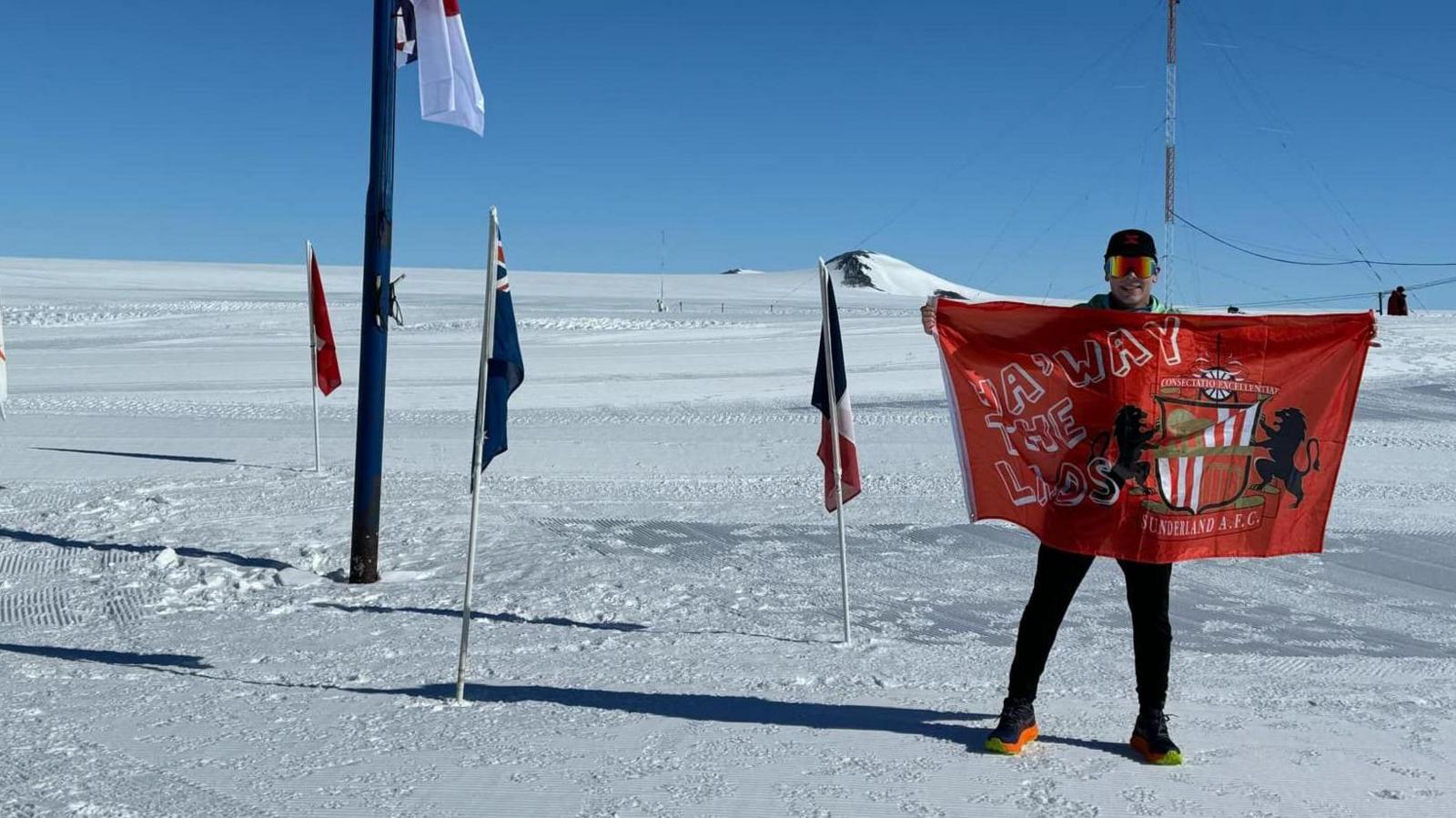 Paul Holborn standing in Antarctica holding a big, red Sunderland AFC flag. He's wearing colourful sunglasses and a cap. The ground and horizon is covered in snow. Various flags stand around him.