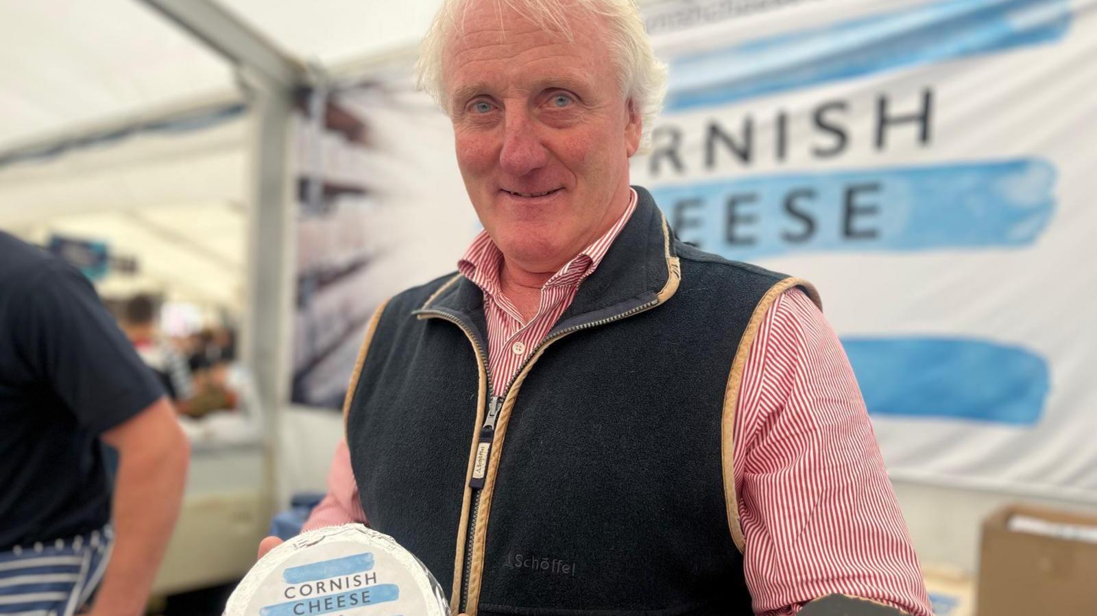 Phillip Stansfield, managing director of the Cornish Cheese Company, holds a wheel of cheese from his company inside a marquee at the Royal Cornwall Show. He is wearing a black gilet and a red and white stripped top.
