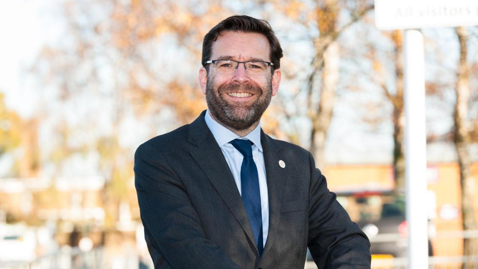 Andy Johnson is smiling at the camera whilst standing outside a school. He's wearing a dark suit on top of a blue shirt and tie.