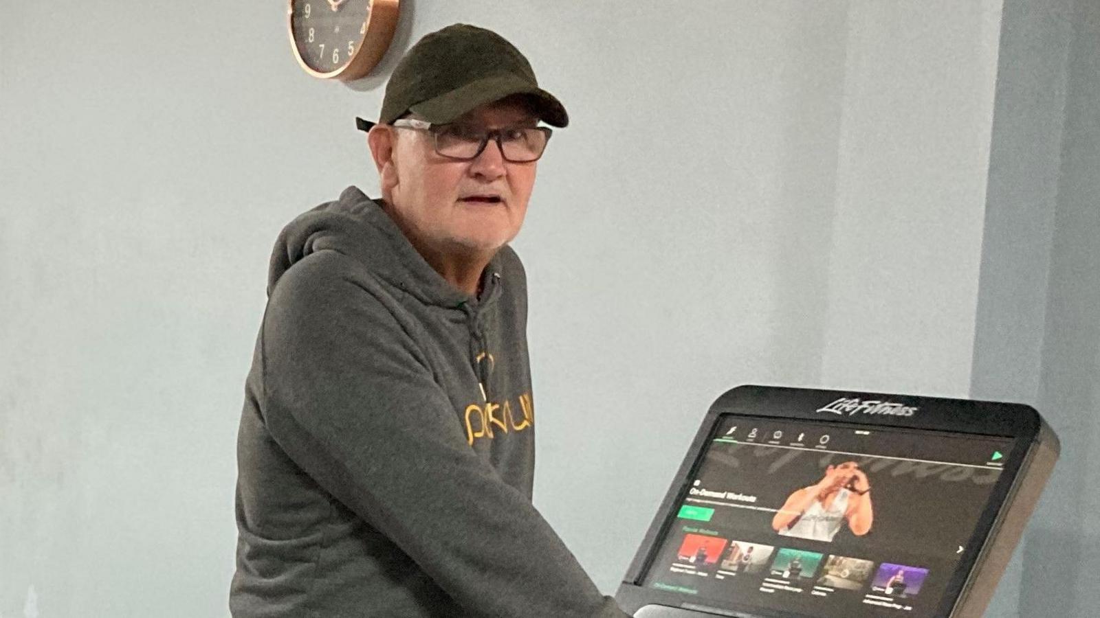 Chris is stood on a treadmill looking at the camera. He is wearing a grey jumper and shorts and a black cap and glasses.